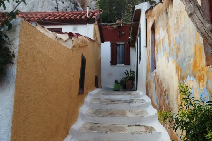 Un callejón de Anafiotika con escalones encalados entre una pared ocre y una casa blanca de contraventanas rojas, bajo la roca de la Acrópolis