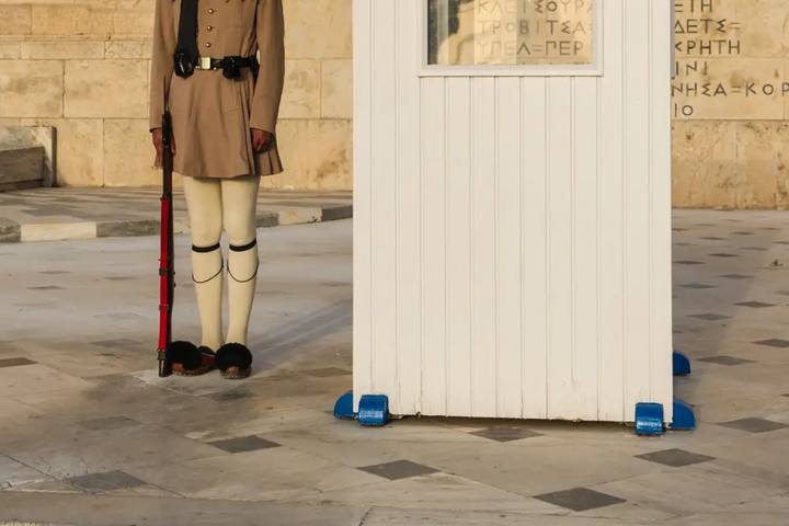 Un evzone de la Guardia Presidencial, de pecho para abajo, con su fusil y sus zapatos de pompón negro, junto a la garita blanca ante el Parlamento