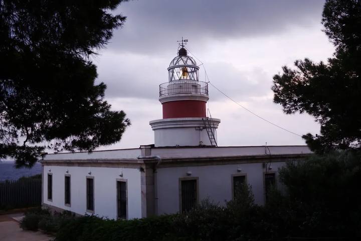 Faro de Salou, torre blanca y roja sobre una casa blanca, enmarcado por pinos en el Cap Salou con cielo nublado